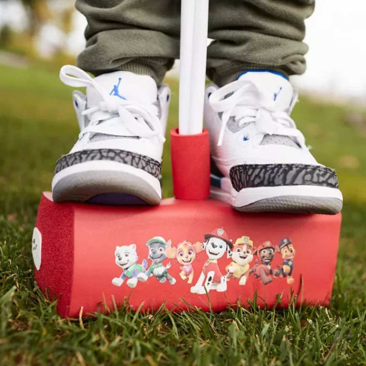 A child in white and gray sneakers stands on grass with a red Paw Patrol Flybar (Marshall) pogo jumper decorated with character stickers.