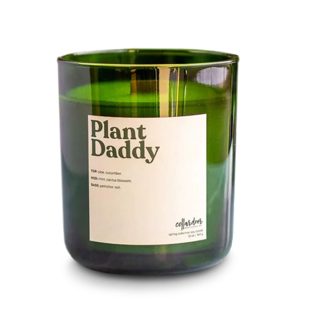 The Plant Daddy Candle is a green glass jar with a white label listing fragrance notes—top: dew, cucumber; mid: mint, cactus blossom; base: patchouli soil. "Cellar Door" is printed at the bottom.