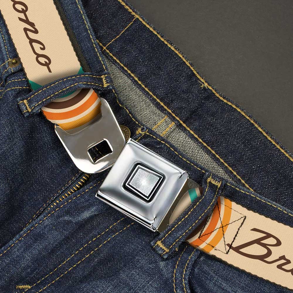 A close-up of blue jeans with the Ford Bronco Script Seatbelt Belt threaded through the loops. This unique automotive accessory is beige with colorful stripes and features “Bronco” printed on it, making it a perfect belt for Ford Bronco enthusiasts.