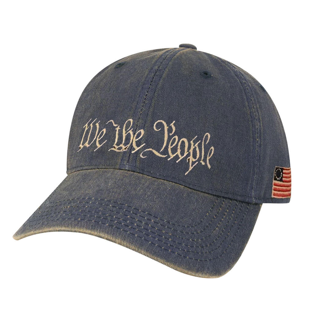 The We the People Cap is a blue denim hat with "We The People" in white script on the front and an American flag patch on the side—ideal for trips to historic sites or expressing your patriotic spirit.