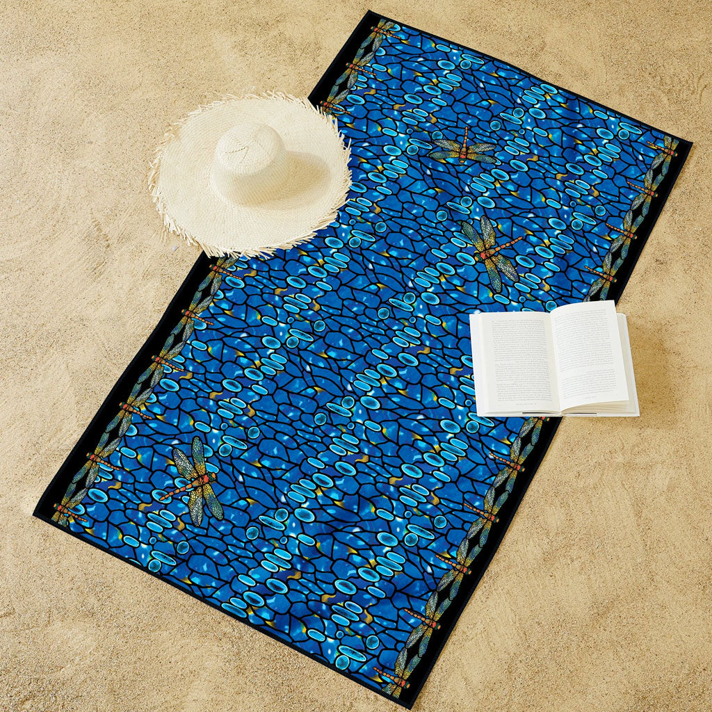 The Louis Comfort Tiffany Dragonfly Beach Towel, featuring blue dragonfly patterns, rests on the sand beside an open book and a straw sun hat with a frayed brim.