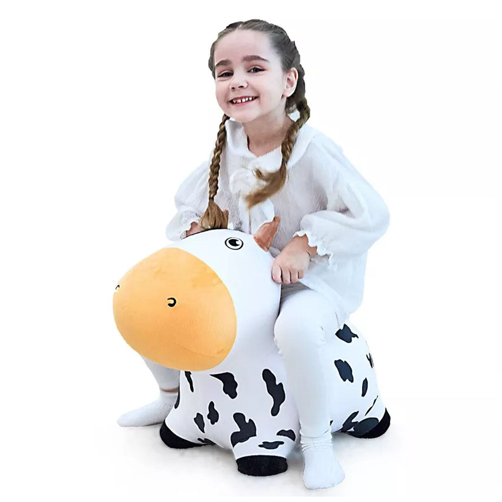 A smiling young girl with braided hair, dressed in white, sits on the Bouncy Pals Cow, a plush black-and-white ride-on toy, against a plain white background.