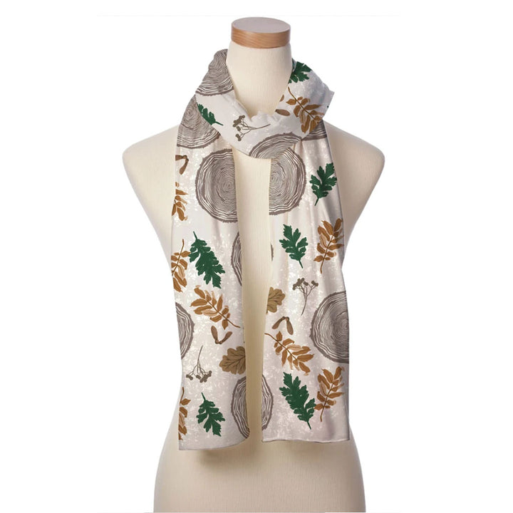 The Wood Tree Silky Scarf, featuring a cream base with brown tree rings and green and gold leaf details, is draped around a beige dress form mannequin.