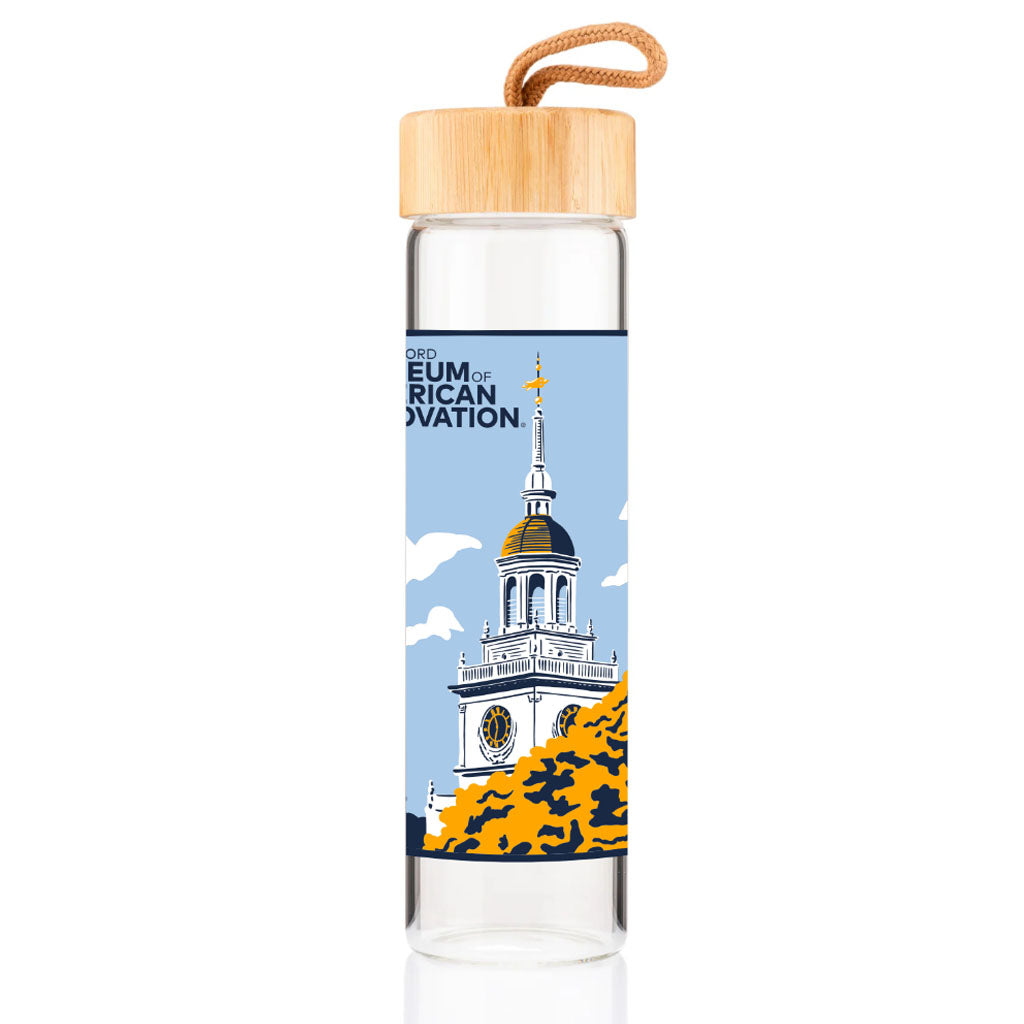 The Henry Ford Museum Building Glass Water Bottle features clear glass, a bamboo lid with strap, and an illustration of the museum’s domed building with clock tower and gold accents, labeled "Museum of American Innovation" on a blue background.