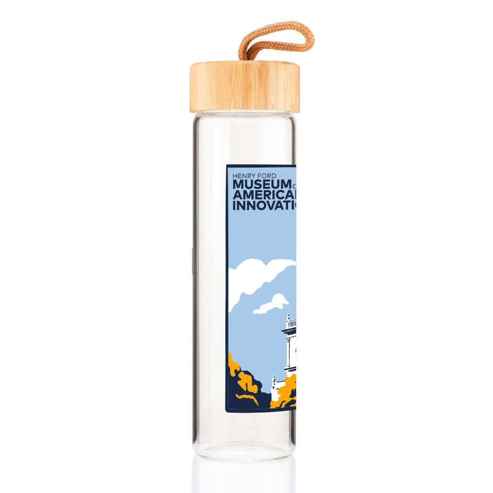 The Henry Ford Museum Building Glass Water Bottle features a clear glass body, bamboo lid, tan carrying loop, and a label with "Henry Ford Museum of American Innovation" plus an illustrated building and trees.