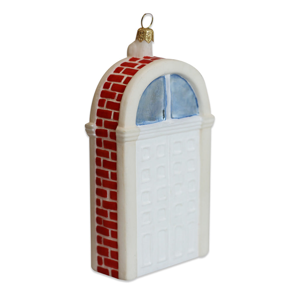 The Henry Ford Museum Festive Doors Ornament is a blown glass arched white door with red brick trim, blue windows, and a gold hanging loop—perfect for adding a unique touch to your holiday décor.