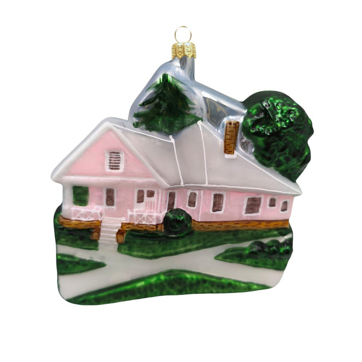 The Henry Ford® Jackson Home Ornament is a glass ornament shaped like a pink house with porch, green bushes, tree, and chimney. Inspired by the Jackson Home at The Henry Ford Museum, it features a gold cap and loop for hanging.