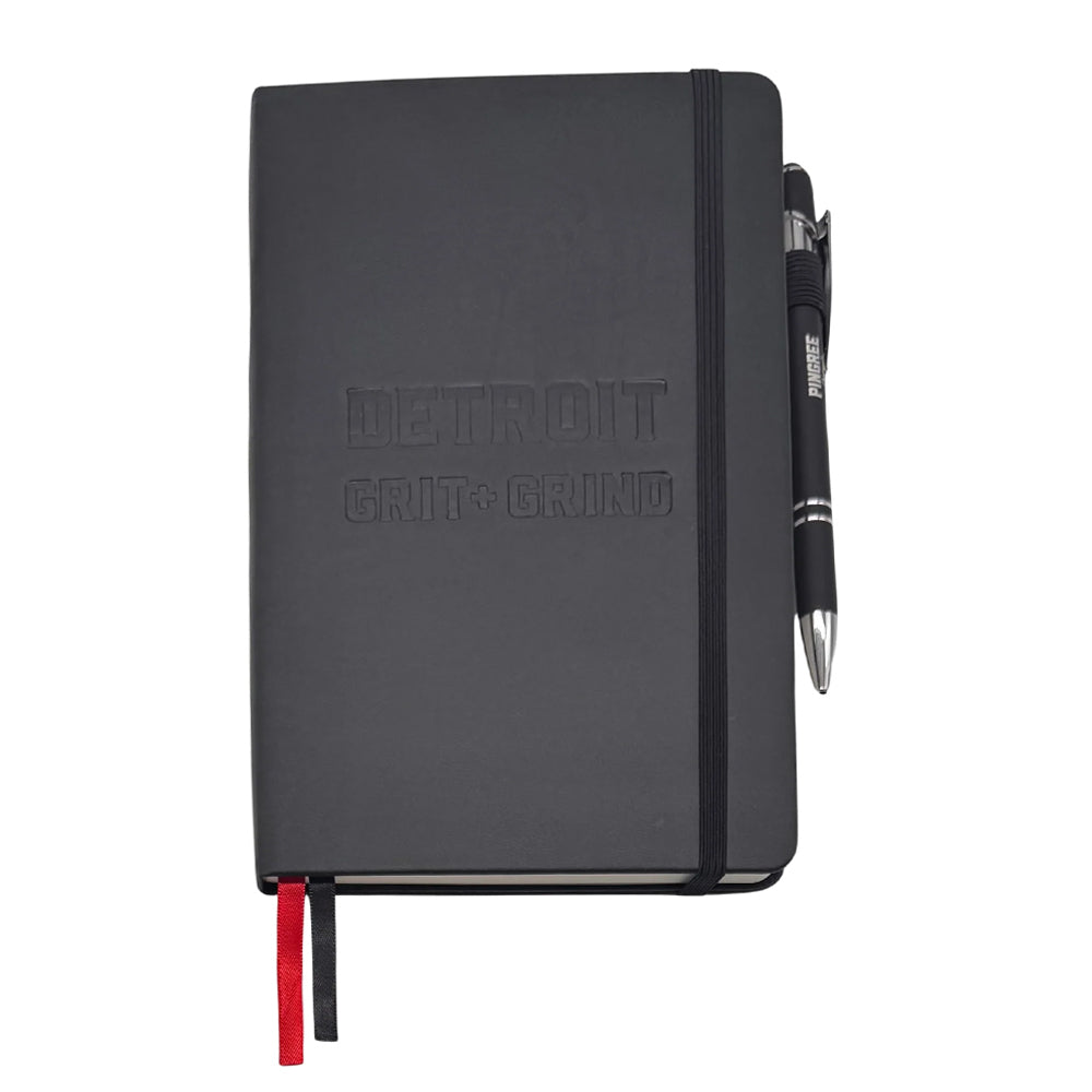 The Detroit Grit and Grind Journal is a black lay-flat journal with "DETROIT GRIT + GRIND" embossed on the cover, an elastic closure band, black pen, and red and black ribbon bookmarks.