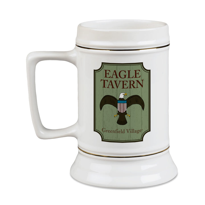 Introducing the Greenfield Village Eagle Tavern Stein—a white ceramic beer stein with a sturdy handle. It features a distinctive label that reads “Eagle Tavern, Greenfield Village” along with an illustrated eagle bearing a shield on its chest. Capturing the historic charm of 19th-century America, this collectible is ideal for history enthusiasts and devoted fans of Greenfield Village.