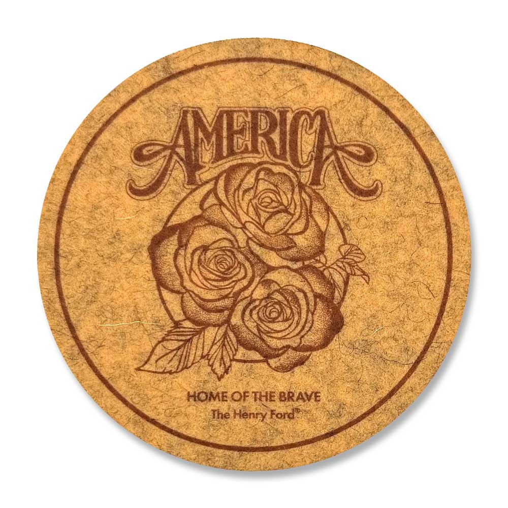 A round, yellow coaster featuring "AMERICA" in stylized text above roses, with "HOME OF THE BRAVE The Henry Ford Museum" below—an ideal keepsake for The Henry Ford® America's 250th Coaster collection.