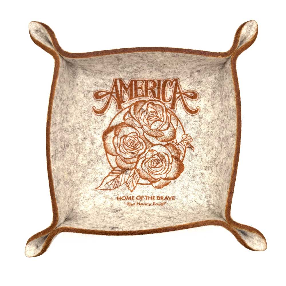 The Henry Ford® America's 250th Trinket Dish is a square felt tray with brown edges, a floral design, “AMERICA” above two roses, and "HOME OF THE BRAVE" below. Made from durable Merino Wool and inspired by the Henry Ford Museum.