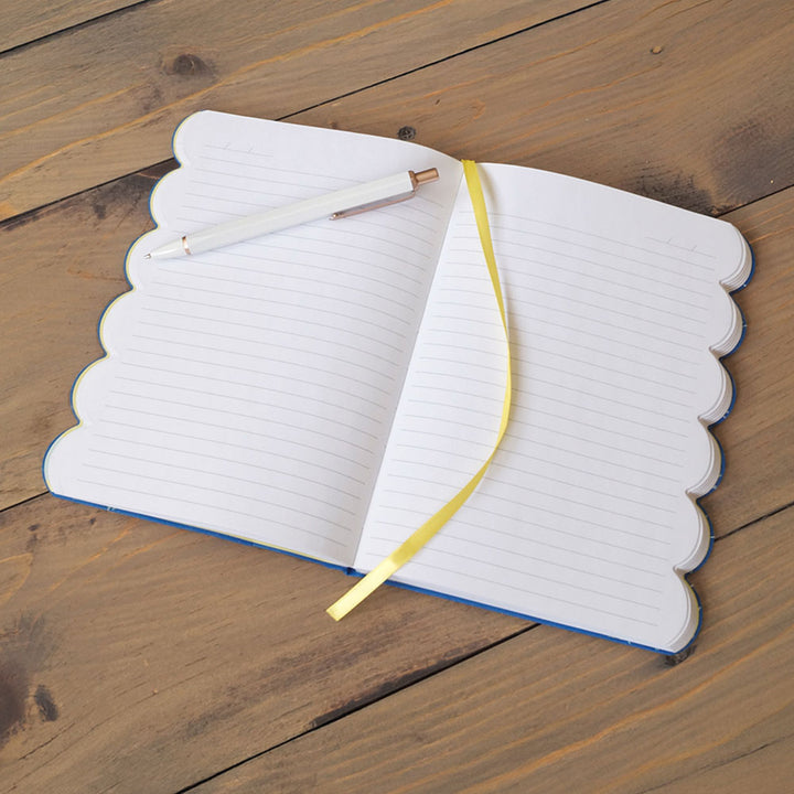 The Eyelet Embroidered Journal, featuring scalloped edges, lined pages, and a yellow ribbon bookmark, rests on a wooden surface with a white pen with a gold tip placed across its left page.
