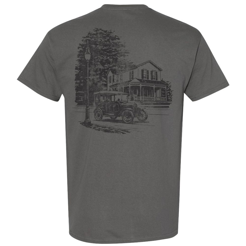 The Greenfield Village Historic Streetscape T-Shirt features a gray design with a black sketch on the back, depicting a vintage car parked beside an old house and streetlamp, framed by trees in the background. Inspired by Greenfield Village, this t-shirt captures the charm of an early 20th-century streetscape reminiscent of a living museum.