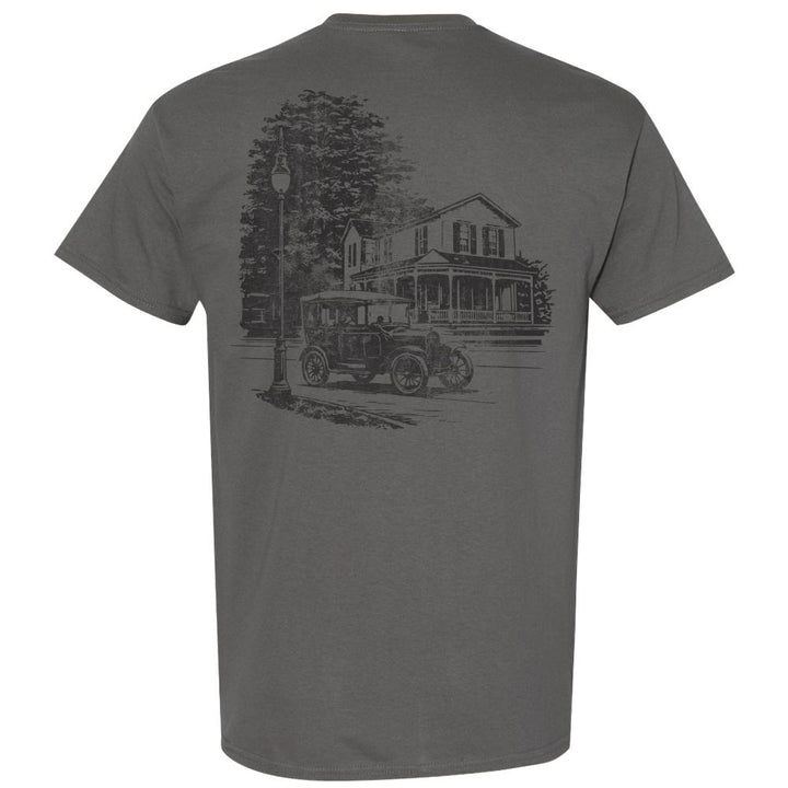 The Greenfield Village Historic Streetscape T-Shirt features a gray design with a black sketch on the back, depicting a vintage car parked beside an old house and streetlamp, framed by trees in the background. Inspired by Greenfield Village, this t-shirt captures the charm of an early 20th-century streetscape reminiscent of a living museum.