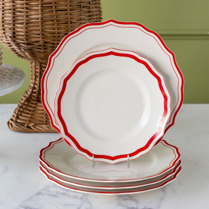 Five Red Icing on the Cake Salad Plates, featuring scalloped edges and a bold red trim, are stacked on a marble surface. Crafted from chip-resistant porcelain, one plate stands upright in a holder while a woven basket rests in the background against a green wall.