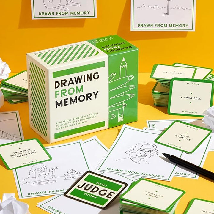 The Drawing From Memory Game features green and white cards, a box, a pen, and scattered drawings on a yellow surface. Some cards display sketches and phrases, with crumpled paper also present.