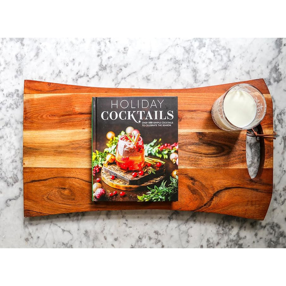 A wooden serving board displays the book "Holiday Cocktails: Over 100 Simple Cocktails to Celebrate the Season" alongside a glass of milk with a cinnamon stick, all arranged on a white marble surface.