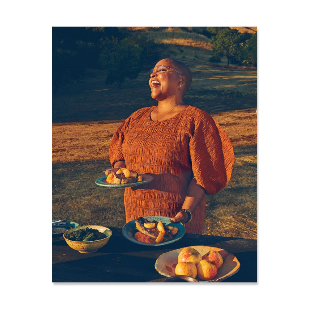A person in an orange dress stands outdoors at a table with plates from "Tanya Holland's California Soul: Recipes from a Culinary Journey West," smiling under warm sunlight amid grassy fields and trees.