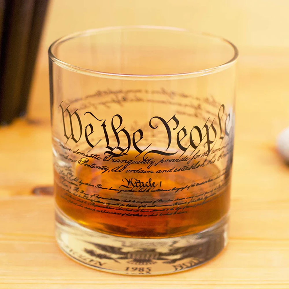 Enjoy your favorite whiskey in the Declaration of Independence Rocks Glass, featuring "We the People" from the U.S. Constitution elegantly printed in black script on a sturdy glass atop a classic wooden surface.
