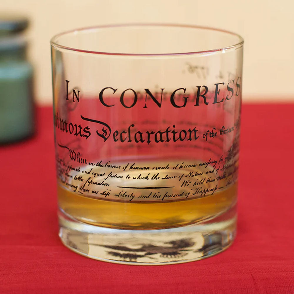 A U.S. Constitution Rocks Glass, partially filled with amber liquid, rests on a red surface and features the historic Constitution text printed on its side for a unique touch.