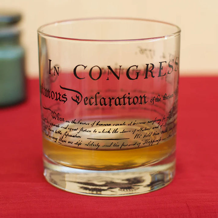 A U.S. Constitution Rocks Glass, partially filled with amber liquid, rests on a red surface and features the historic Constitution text printed on its side for a unique touch.
