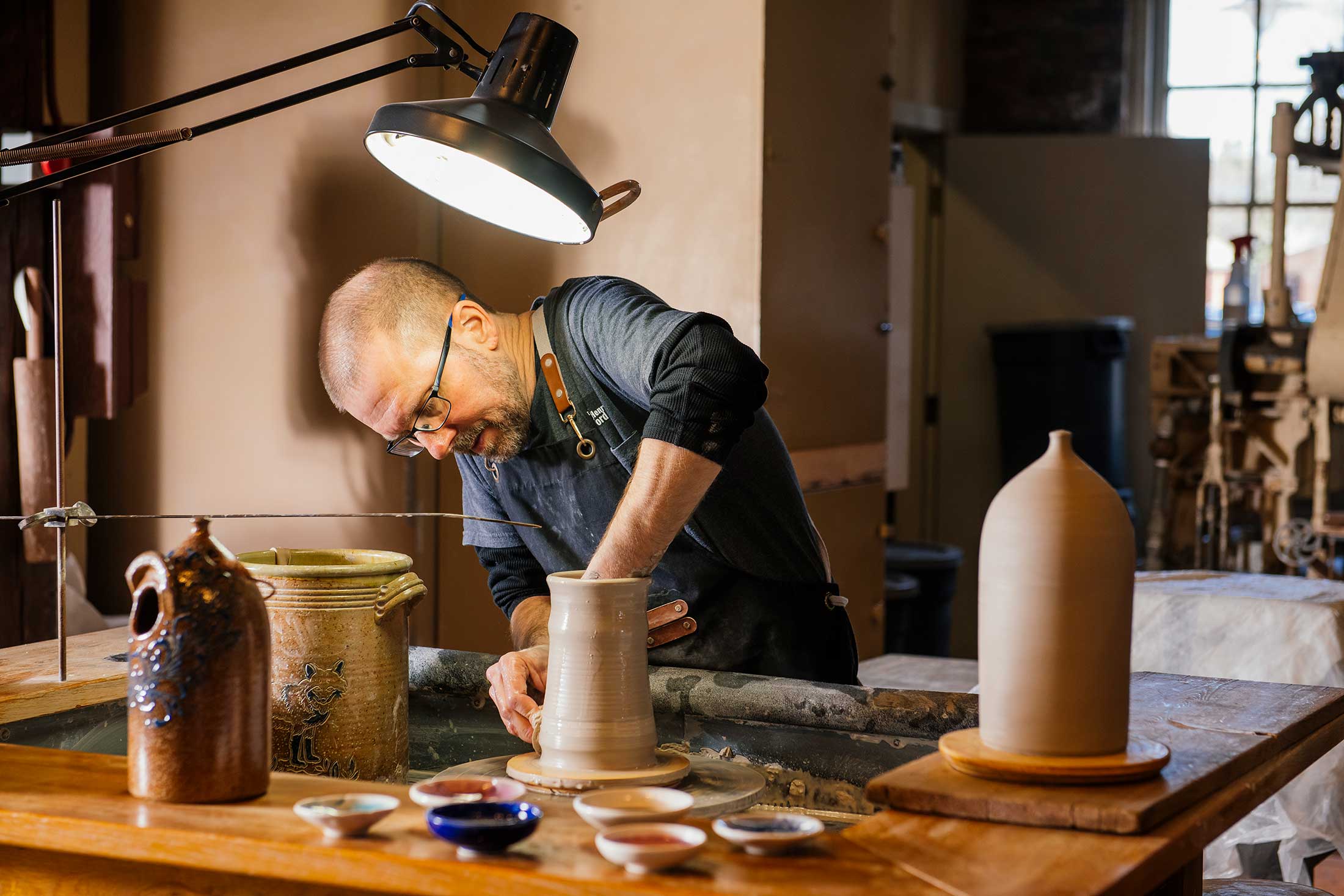 A man wearing glasses and an apron shapes clay on a pottery wheel in a well-lit studio, surrounded by finished pottery pieces and small bowls on a wooden table.