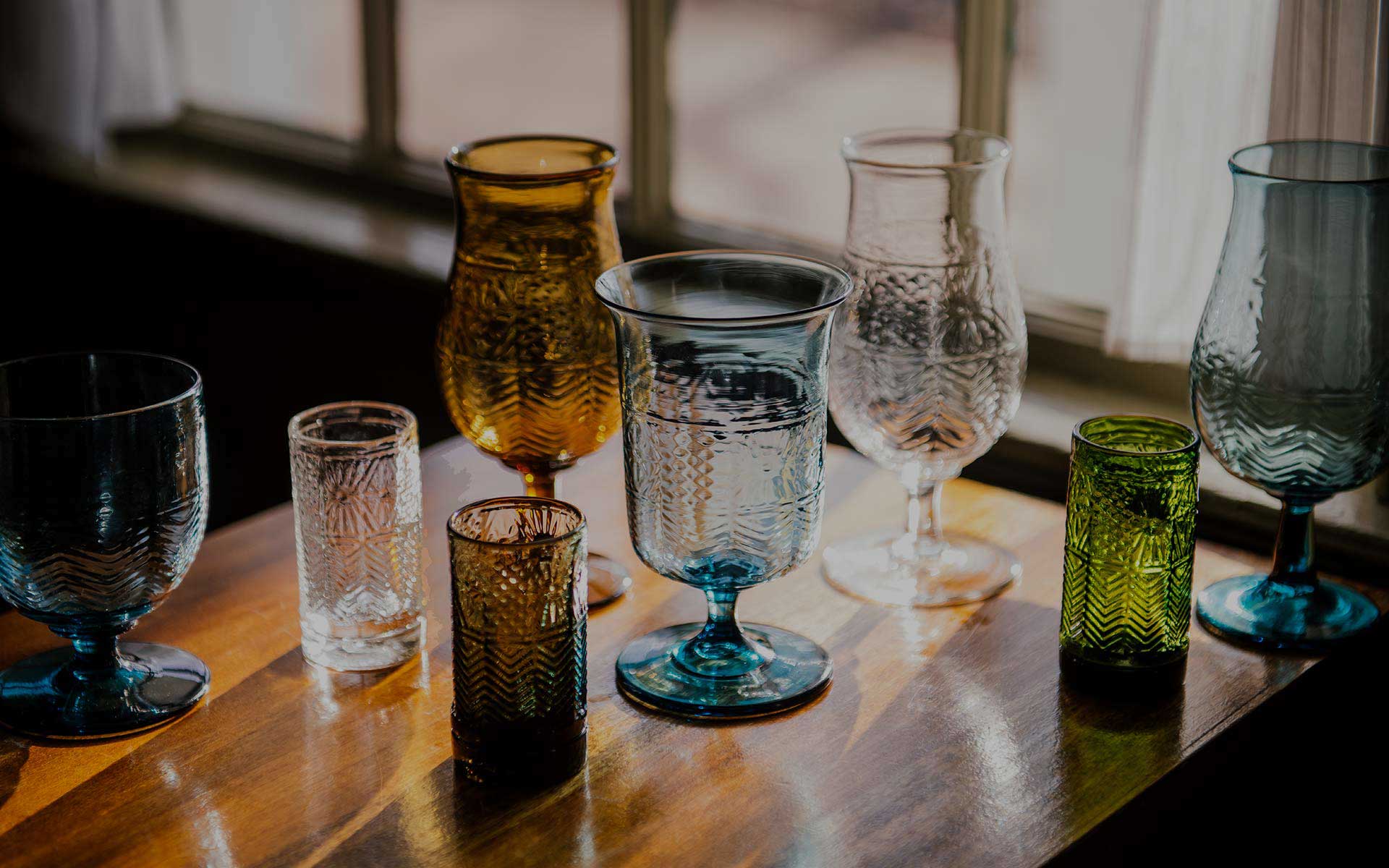 A variety of colorful textured glass cups and goblets sit on a wooden table by a sunlit window, casting soft shadows and light reflections.