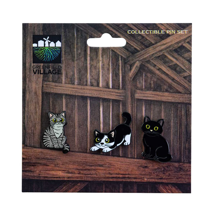 Three cartoon cat pins—one gray tabby, one black and white, and one solid black—are displayed on a card with a barn wood background labeled “Greenfield Village Barn Cats Pin Set” at the top left, celebrating the feline residents of Greenfield Village.