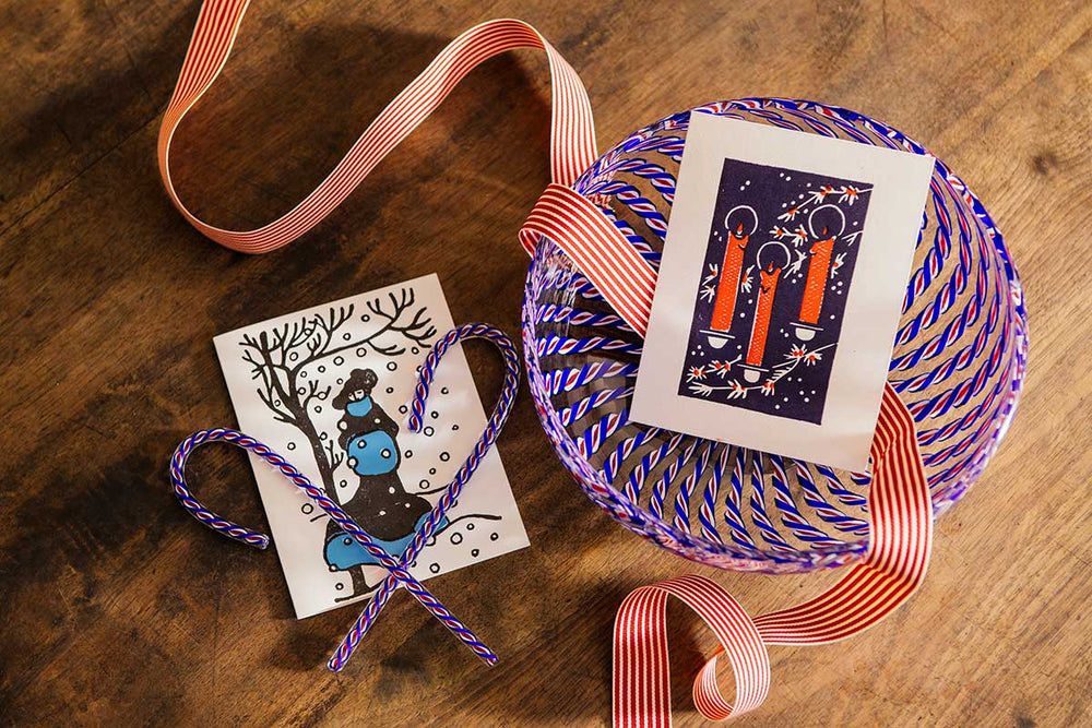 The 2026 Candy Cane Card Set, featuring a red candle card and a winter deer scene with a glass candy cane accent, is displayed alongside a bowl tied with a red-and-white striped ribbon on a wooden surface.
