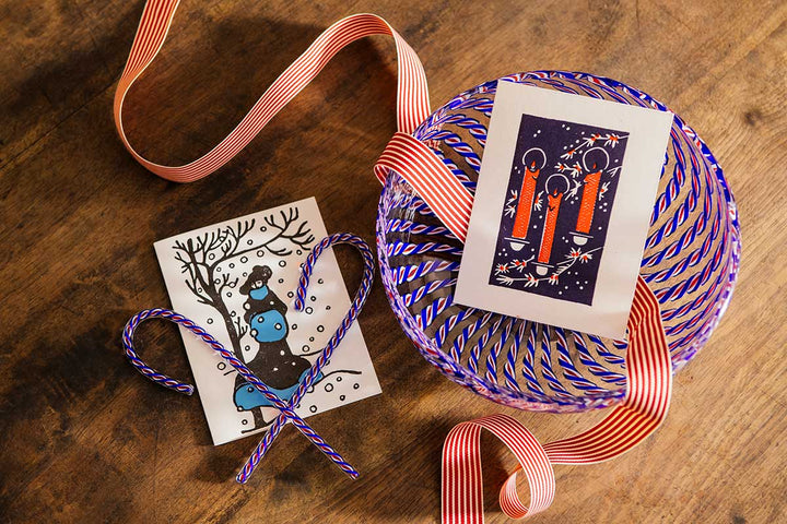 The 2026 Candy Cane Card Set, featuring a red candle card and a winter deer scene with a glass candy cane accent, is displayed alongside a bowl tied with a red-and-white striped ribbon on a wooden surface.