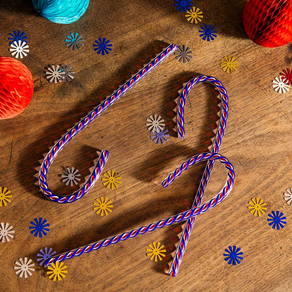 Three 2026 Candy Cane limited editions with blue, white, and red stripes rest on a wooden surface sprinkled with colorful confetti and honeycomb paper balls in red, orange, and turquoise.