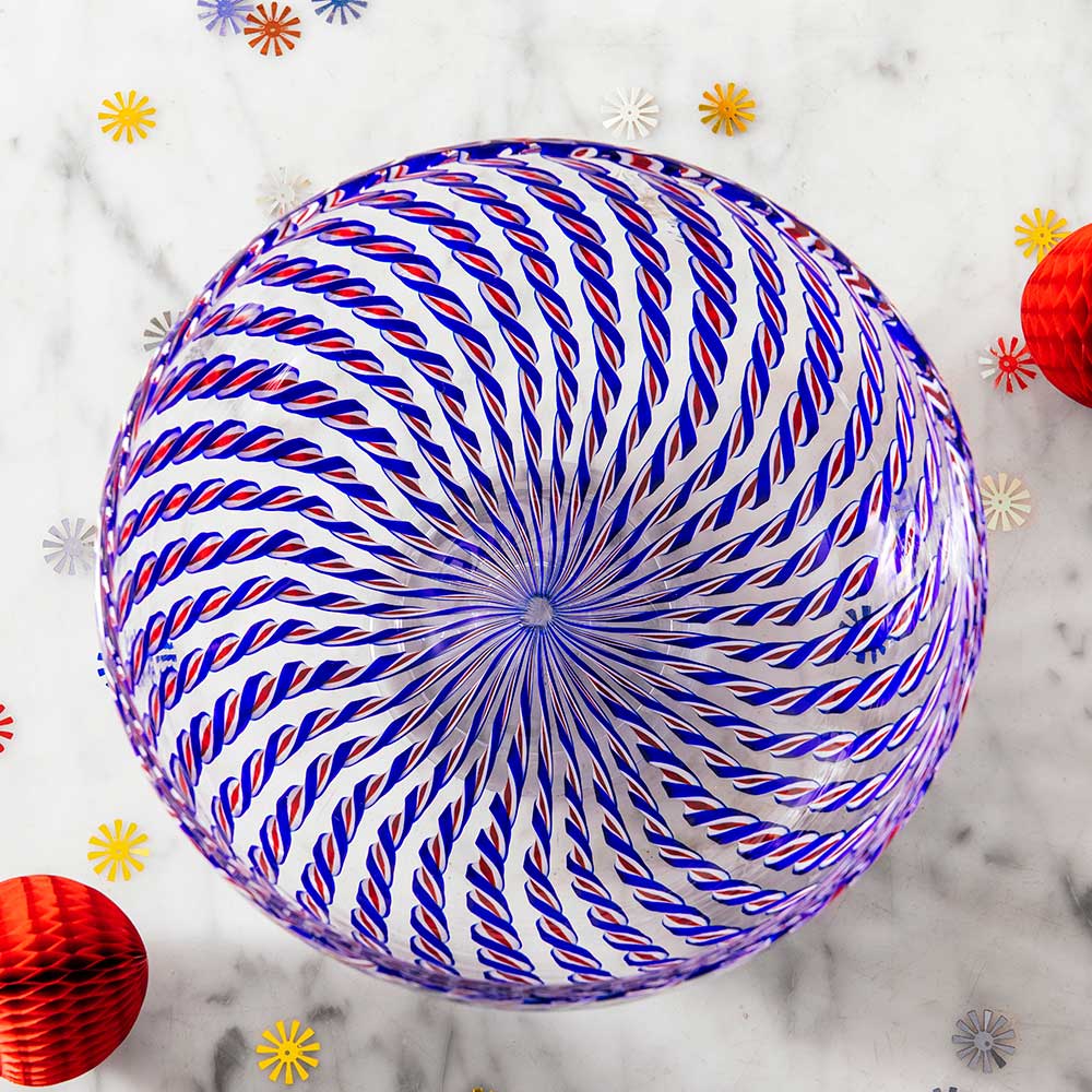 The 2026 Candy Cane Bowl, featuring blue and red swirling patterns, sits on a marble surface with yellow and red decorative balls and flower-shaped confetti, showcasing the artistry of America's 250th glassblowers.