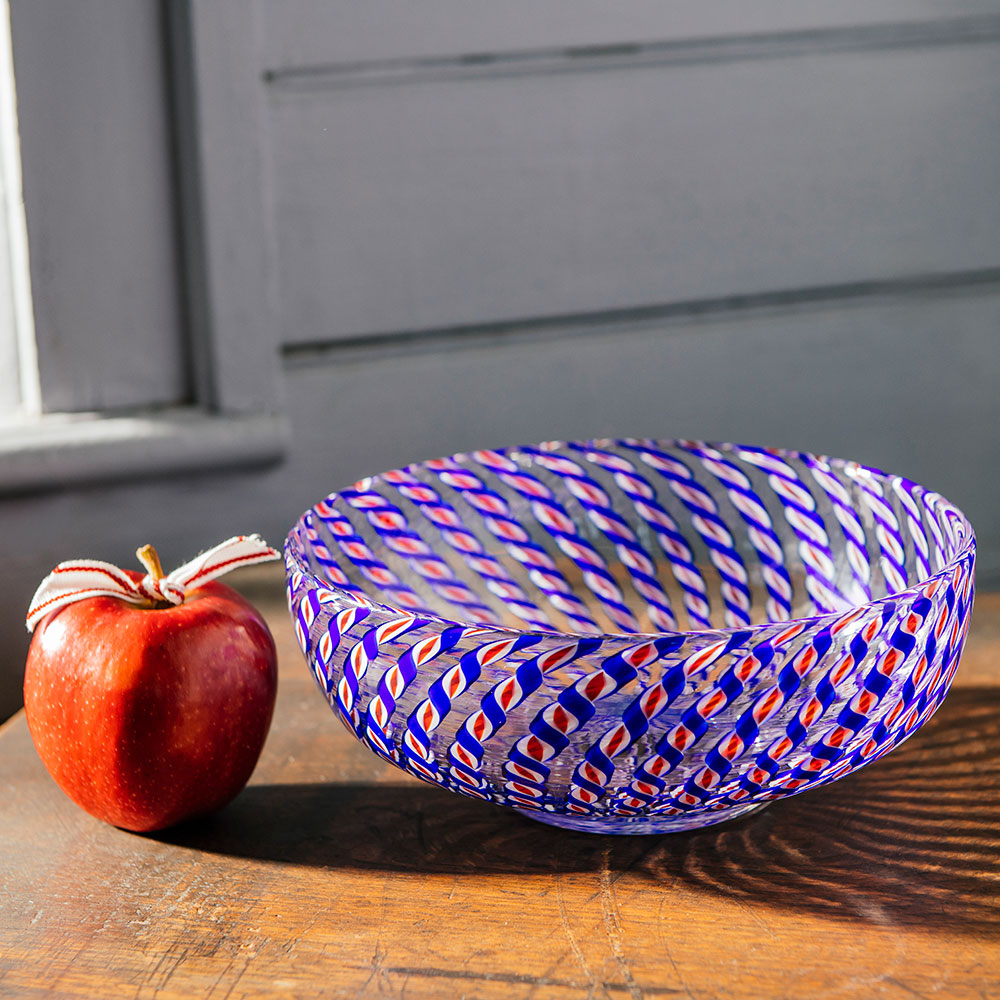 A red apple with a decorative ribbon sits next to the 2026 Candy Cane Bowl, crafted by skilled glassblowers and featuring swirling blue, red, and white patterns, on a wooden table in front of a gray wall.