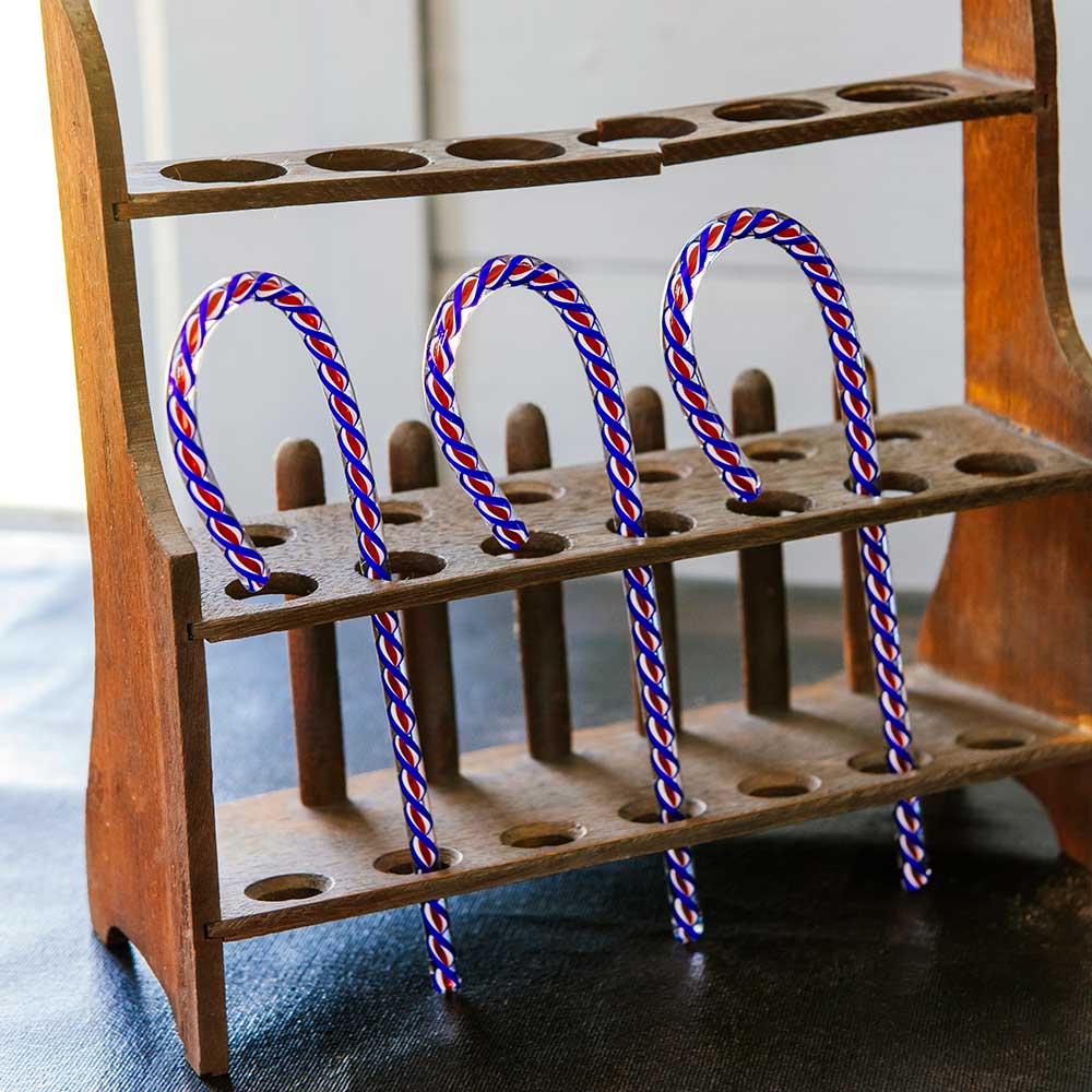 Four 2026 Candy Cane limited-edition red, white, and blue striped candy canes are arranged in a wooden test tube rack, their curved ends sticking up—a festive way to honor America’s 250th.