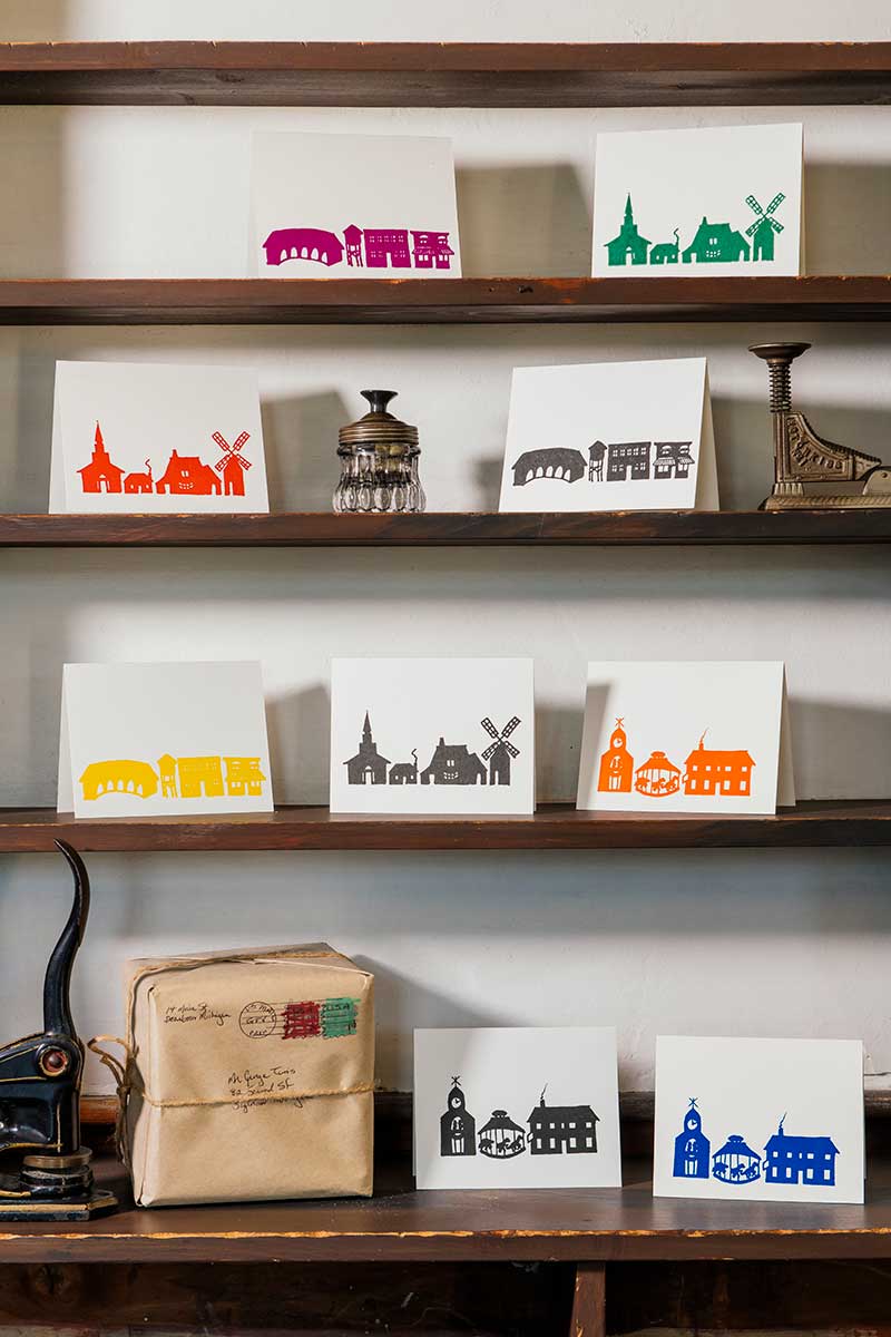 The Rainbow Village Card Set of 6, featuring colorful silhouettes of Greenfield Village landmarks, is displayed on wooden shelves with vintage items. A wrapped parcel and two more cards rest on the lower shelf.
