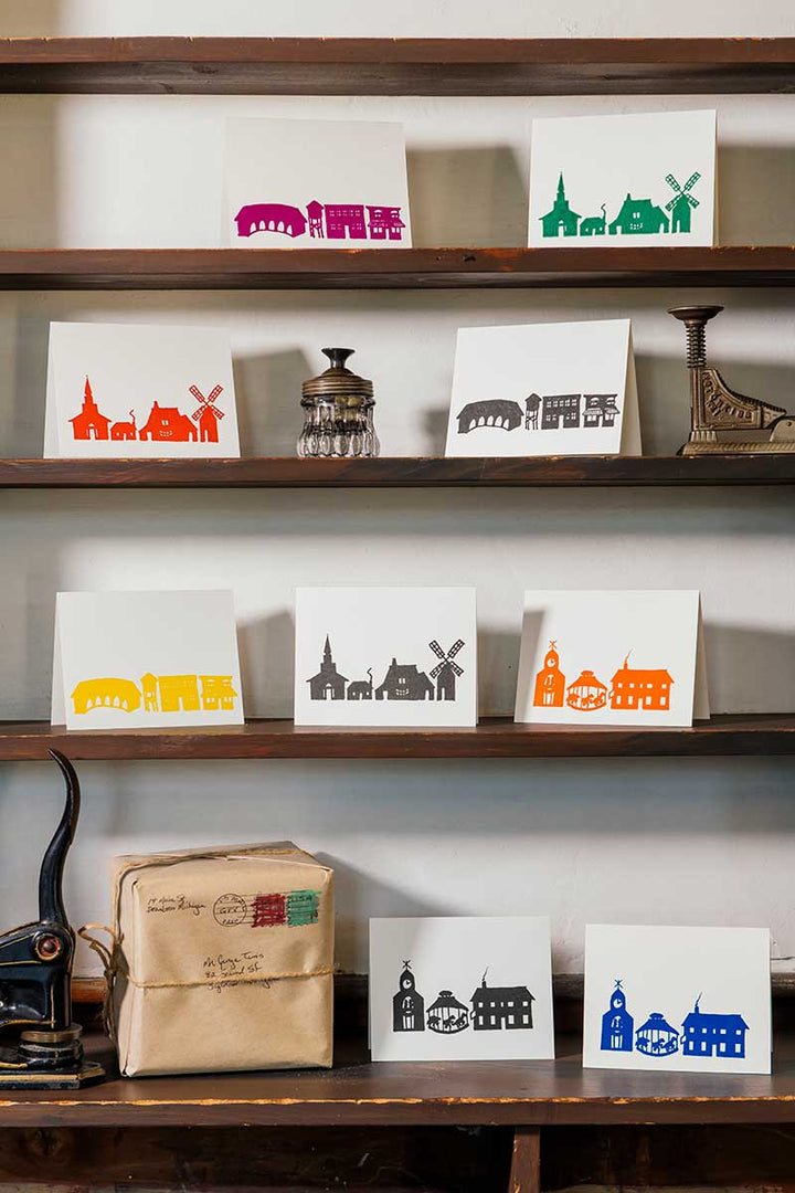 The Rainbow Village Card Set of 6, featuring colorful silhouettes of Greenfield Village landmarks, is displayed on wooden shelves with vintage items. A wrapped parcel and two more cards rest on the lower shelf.