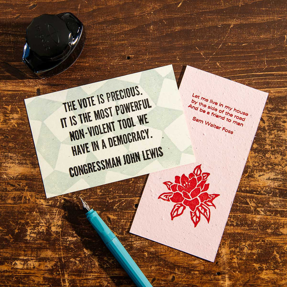 Two cards rest on a wooden table: one features an inspirational excerpt from a John Lewis quote about voting, while the other displays a red flower alongside a Sam Walter Foss quote. Also visible are the House by the Side of the Road Bookmark, a teal pen, and a black ink bottle.