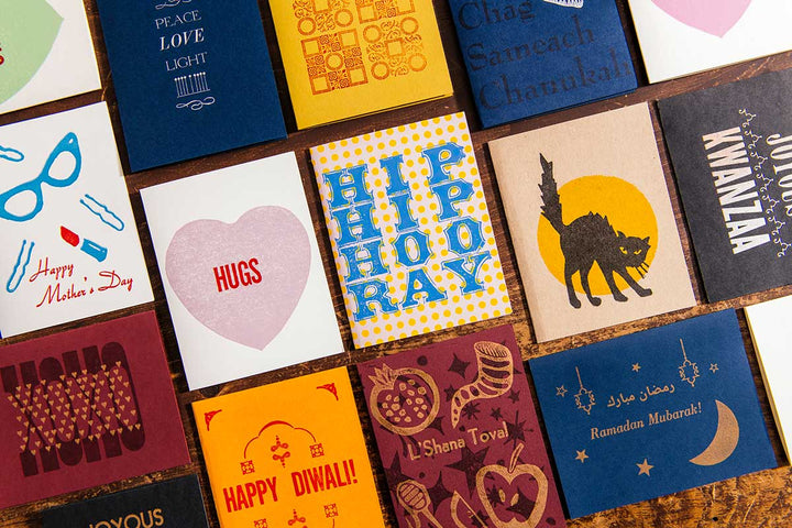 A handmade Kwanzaa Card on black paper is displayed with other colorful greeting cards for occasions like Mother’s Day, Hanukkah, Diwali, Ramadan, and more, all arranged in a grid on a wooden surface.