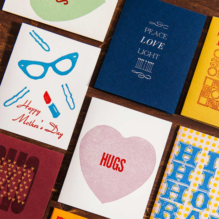 A colorful handcrafted Mother's Day Card from Liberty Craftworks sits on a wooden surface, with designs and messages like “Happy Mother’s Day,” “HUGS,” and “PEACE LOVE LIGHT.”.