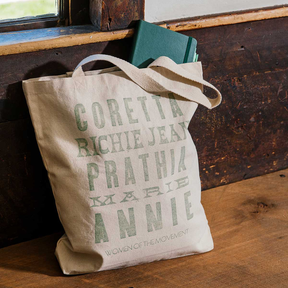 The Women of the Movement Tote, a beige cotton canvas bag featuring green text with names like Coretta, Richie Jean, Prathia, Marie, and Annie, sits on a wooden surface with a book partially visible inside. This handcrafted tote honors influential women of the Civil Rights Movement.