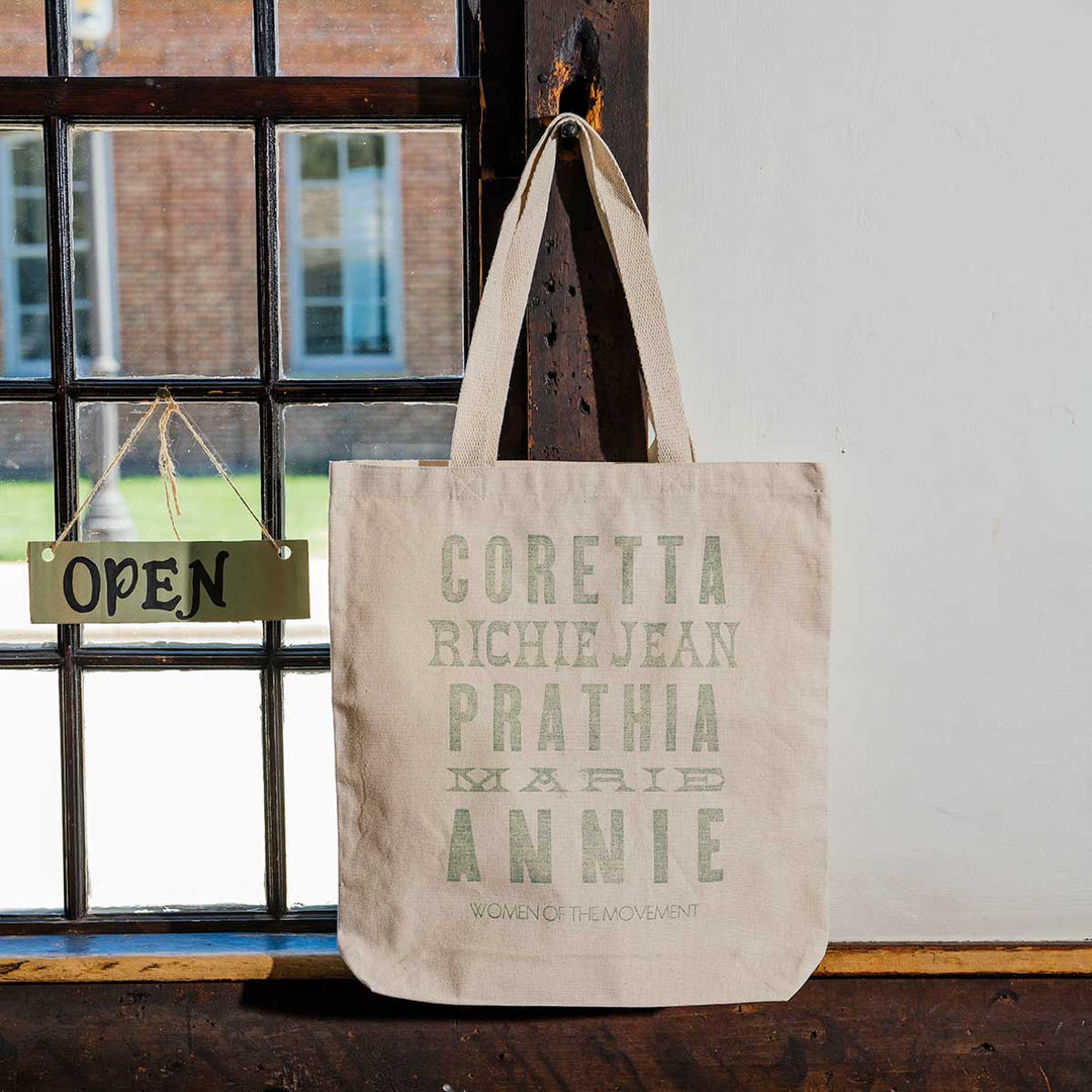 A Women of the Movement Tote, a beige handcrafted cotton canvas bag with names printed on it, hangs on a wall near a window. A small OPEN sign is displayed on the window latch as sunlight streams through the panes.