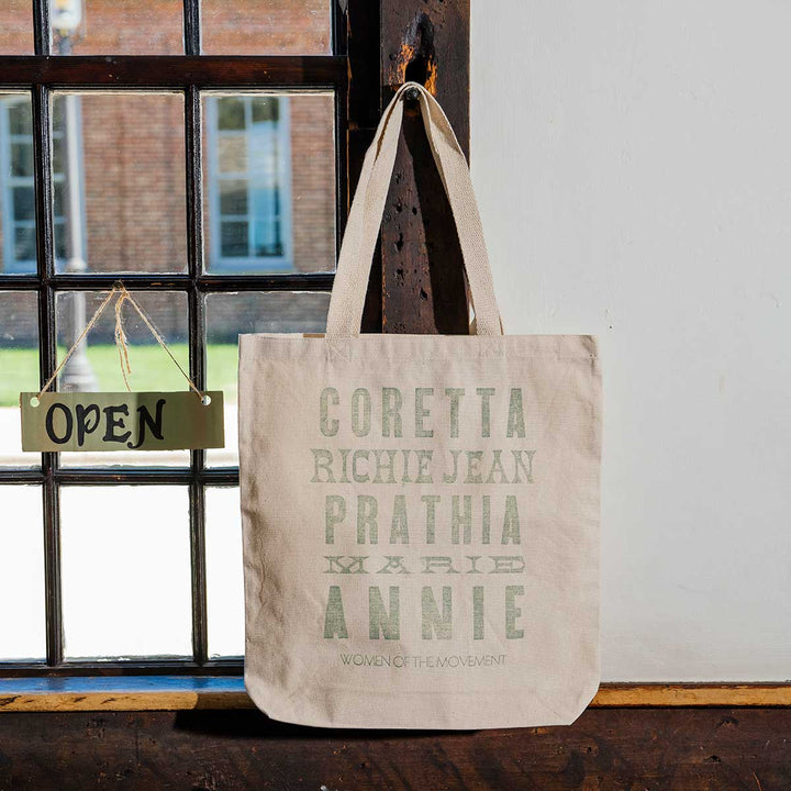 A Women of the Movement Tote, a beige handcrafted cotton canvas bag with names printed on it, hangs on a wall near a window. A small OPEN sign is displayed on the window latch as sunlight streams through the panes.