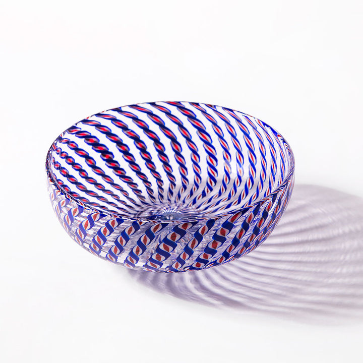 The 2026 Candy Cane Bowl, expertly crafted by glassblowers, features blue, red, and white twisted stripes and casts a striking patterned shadow on a white surface.