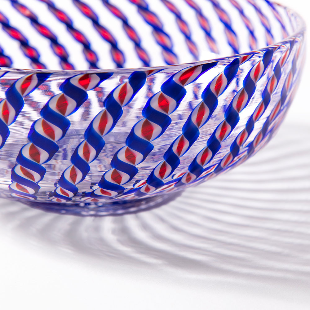 A close-up of the 2026 Candy Cane Bowl, featuring swirling blue and red spiral patterns crafted by skilled glassblowers, casting intricate shadows on a white surface.