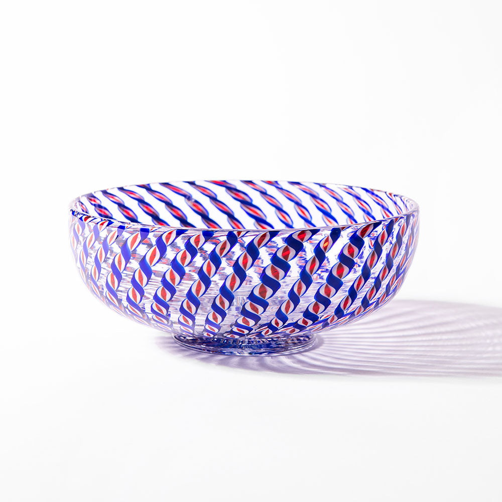 The 2026 Candy Cane Bowl, crafted by skilled glassblowers for America's 250th, features swirling red, blue, and white stripes and casts a vibrant shadow on the surface beneath it.