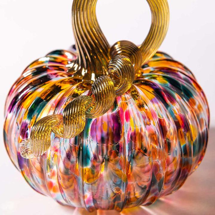 A vibrant Small Glass Pumpkin – Kaleidoscope, hand-blown with swirling patterns of orange, pink, green, and blue, and finished with a shiny gold twisted stem—a delightful collectible inspired by the artistry of Greenfield Village.