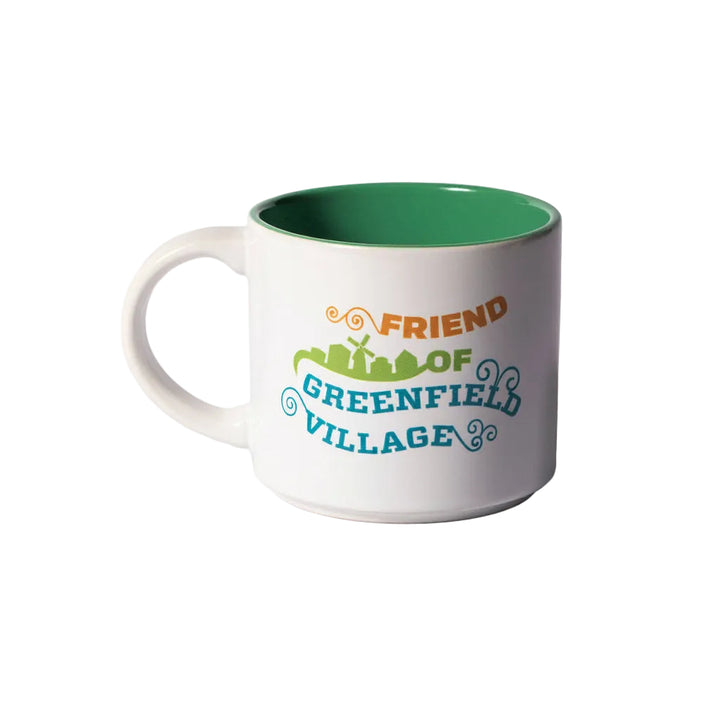 The Friend of Greenfield Village Mug is a white ceramic mug with a green interior, featuring vibrant text that reads “Friend of Greenfield Village” and a graphic depicting buildings and trees inspired by the living history museum. Set against a plain white background, this mug captures the essence of historic American artifacts.