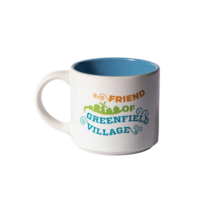 The Friend of Greenfield Village Mug is a white mug featuring a blue interior and colorful text—Friend of Greenfield Village—alongside a green village silhouette and decorative swirls, capturing the spirit of this renowned living history museum.