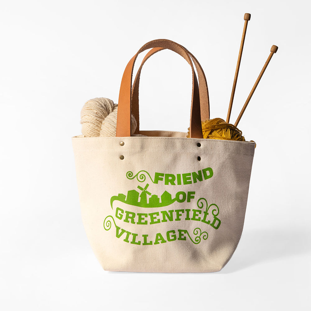The Friend of Greenfield Village Project Tote is a beige canvas bag with brown handles, featuring a green village silhouette and "Friend of Greenfield Village" text, perfect for carrying yarn and knitting needles.