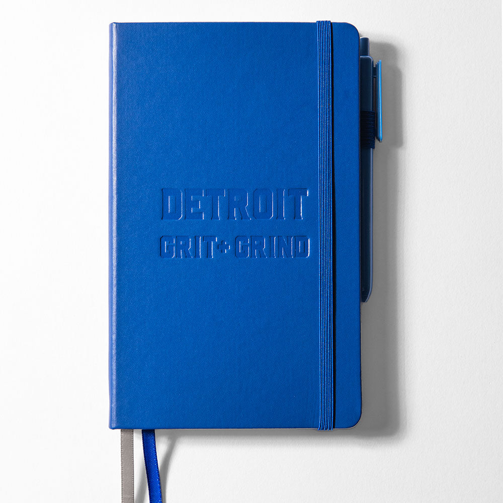 Meet your new favorite Detroit Grit and Grind Journal - Blue: made from recycled paper with DETROIT GRIT + GRIND embossed on the cover, elastic closure band, two ribbon bookmarks, and a pen attached on the right.